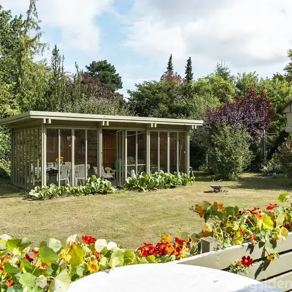 En havepavillon står ubenyttet i baghaven. Pavillonen er omgivet af grønne buske og træer, mens blomster pryder forgrunden. Et fuglehus er placeret på en stolpe til højre.