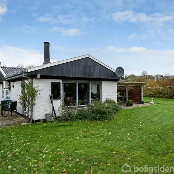 Et lille, hvidt hus med sort tag står i en frodig have. Skorstenen peger opad, og en terrasse med siddepladser ses til venstre omgivet af grønne buske og træer.
