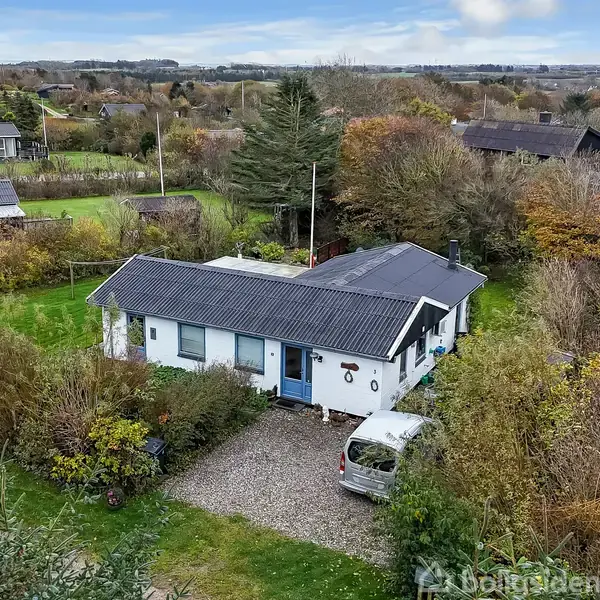 Et hvidt hus med sort tag står stille omgivet af frodig have. En bil er parkeret på grusindkørslen. Landskabet består af grønne marker og flere små huse.