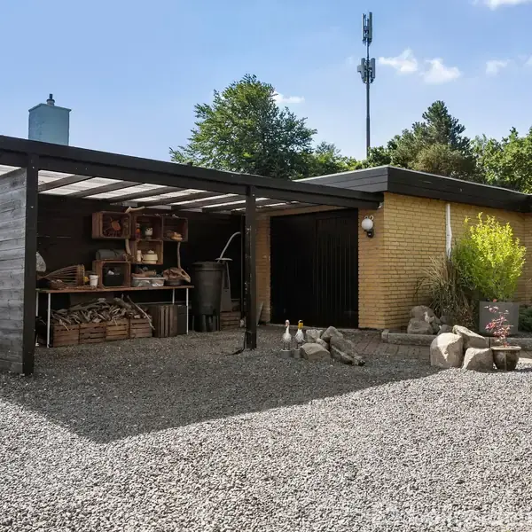 Carport med trætag opbevarer brændestabler og værktøj i en have med grus og mange planter. Det gule murstenshus og blå himmel skaber en harmonisk udendørsscene.
