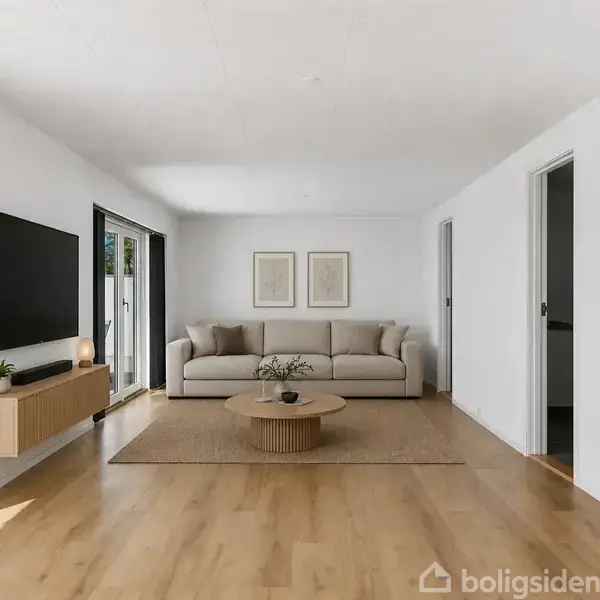 En beige sofa står foran en hvid væg med to billeder. Der er et rundt sofabord foran sofaen. Til venstre hænger et TV på væggen over en skænk.
