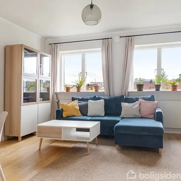 Stue med en blå hjørnesofa, pyntepuder og sofabord. Rummet har store vinduer med gardiner og planter, og en reol samt tv-møbel. Et tilstødende kontor ses til venstre.