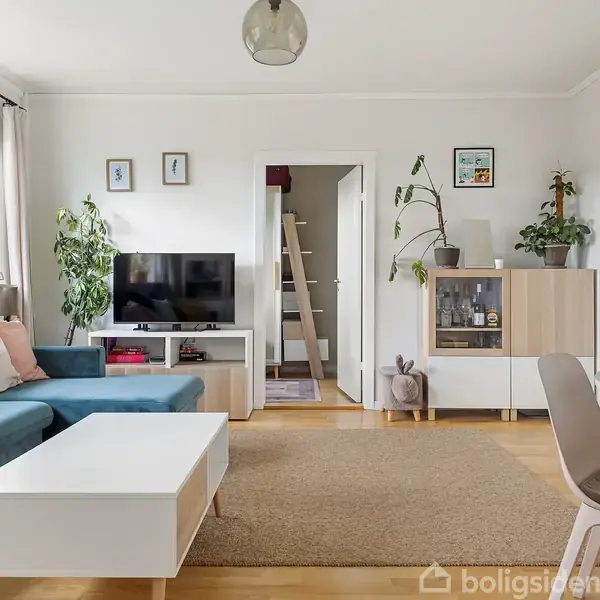 En stue med en blå sofa, sofabord og TV på en TV-bænk. Væggene har billeder, og der er planter på skænken. Bord med stole står i højre hjørne.