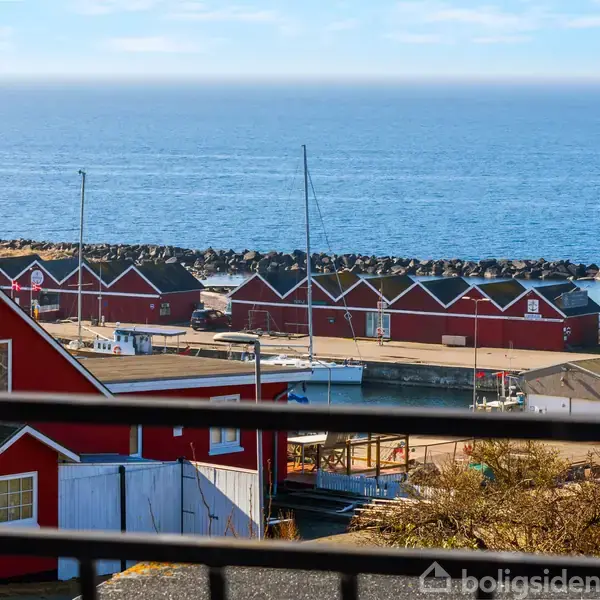 Røde huse langs kysten står stille. Havet breder sig ud i baggrunden med en stenmole, der beskytter mod bølgerne, under en blå himmel.