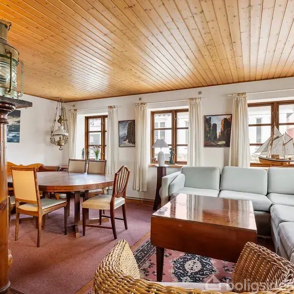 Stue med en stor lysegrå hjørnesofa og et træbord. Omkring står træstole ved et rundt spisebord. Væggene har maritime dekorationer og vinduer med lyse gardiner giver lys.