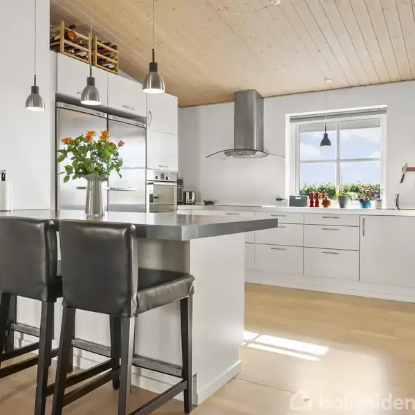 Et moderne køkken med hvidt skab og køkkenbord, tre sorte barstole, og blomster i en vase. Der er opvaskemaskine, køkkenredskaber på væggen, og solen lyser gennem et vindue.