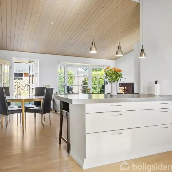 Et moderne køkken-alrum med en hvid køkkenø fyldt med orange og gule blomster. Alrummet har lyse træmøbler, blå vægge, træloft og et spisebord ved vinduet, der slipper naturligt lys ind.