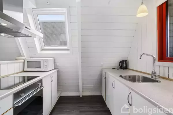 Et hvidt køkken med en mikrobølgeovn og ovn på en bordplade, en vandhane ved vasken. Et skråt vindue og rødt indrammet vindue lader lys ind i rummet.