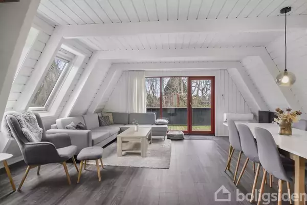 Stue med lyse grå møbler arrangeret omkring et sofabord, i en skråvægget, hvidmalet loftslejlighed. Store vinduer og glasdør leder ud til balkon med udsigt til træer.