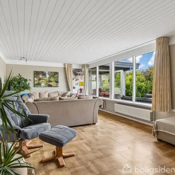 Stue med sofa og lænestol, placerer sig ved panoramavinduer, som slipper lys ind og viser ud til en frodig have. Væggene prydes af kunst og fotografier.