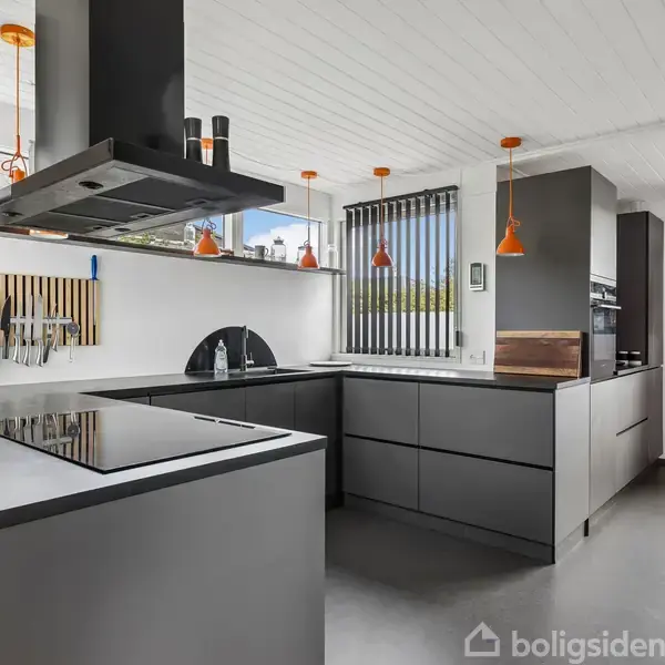 Køkken med grå skabe og sorte bordplader. Lyse indretningselementer inkluderer orange lamper over øen. Store vinduer med persienner slipper naturligt lys ind. Moderne og minimalistisk design.
