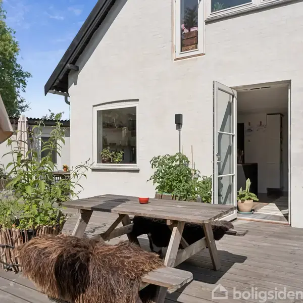 Et træbord med bænke står på en træterrasse. Døren til huset er åben, planter ses i krukker omkring bordet og en parasol.
