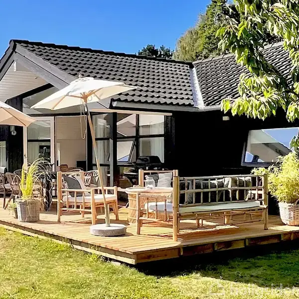 Hus med mørke vægge og sorte tagsten står på en solrig terrasse. Havemøbler og parasoller skaber en afslappet atmosfære omgivet af grønne omgivelser og græsplæne.