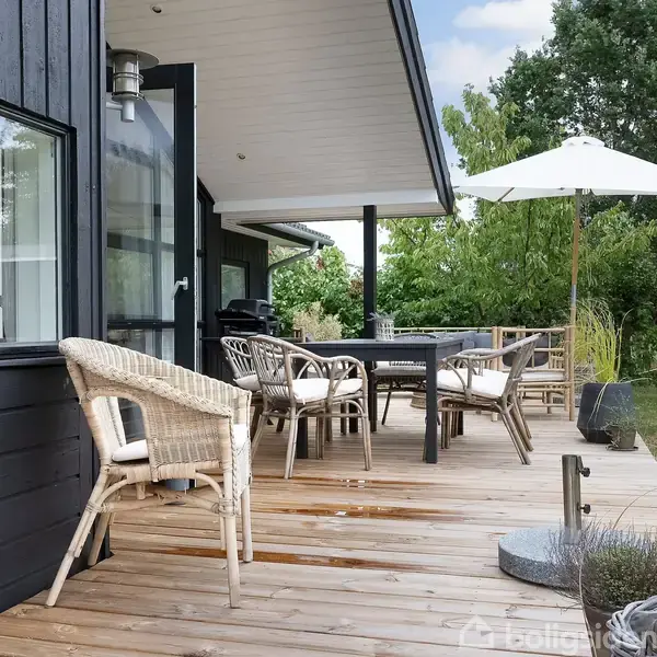 Træterrasse med flettede stole og et bord, omgivet af grønne planter og træer. En stor hvid parasol står ved siden af bordet, under en blå himmel.