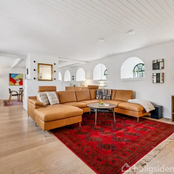 En brun hjørnesofa står på et rødt tæppe i en stue med lyse trægulve og hvide vægge. Vinduer med buet form lader lys ind. Bagved er der et spiseområde.