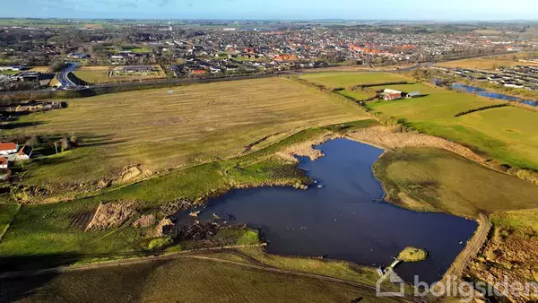 Sø i landskab med græsarealer omgivet af marker og en by i baggrunden. Spredte bygninger ses langs horisonten, og en snoet vej skærer gennem området.
