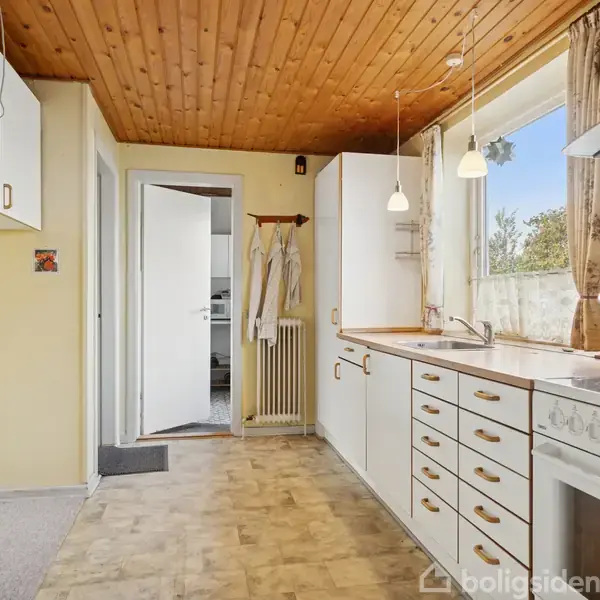 Et køkken med hvide skabe og en ovn til højre. Der er en vask under et stort vindue med udsigt til natur. Lyse trælofter og gulvtæppe.