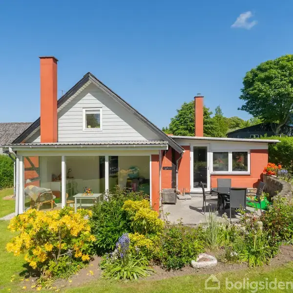 Et mindre hus med orange facade og skråt tag har en veranda med havemøbler. Foran er en farverig have med blomster og buske under en klar blå himmel.
