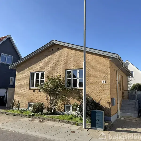 En gul murstenshus står i et beboelseskvarter med en lille have foran. Husnummeret "12" er synligt ved indgangen, og der er en lygtepæl foran huset.