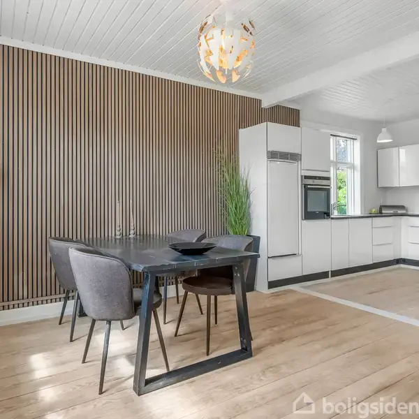 En moderne spisestue og køkken med et sort bord og grå stole. På væggen ses lodrette træpaneler. Køkkenet har hvide skabe og en loftslampe hænger over bordet.