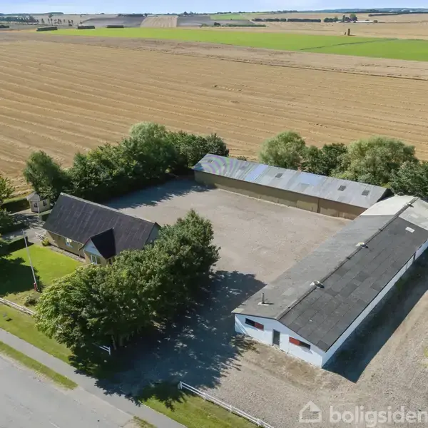 Gårdbygninger omgivet af marker og træer. En hovedbygning og længe danner en grusbelagt gårdsplads. Landlig beliggenhed med åbne marker i baggrunden. Ingen synlig tekst.