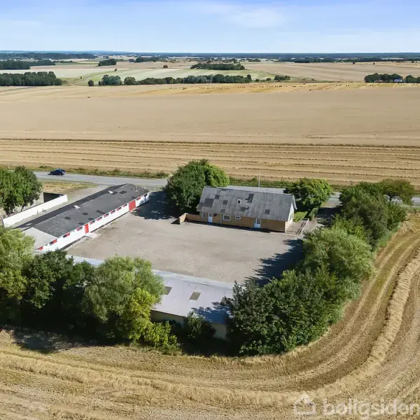 En gårdbygning med en stor gårdsplads omgivet af træer. Bygningen ligger midt i et åbent, høstet marklandskab. En vej ses i baggrunden ved siden af marker.