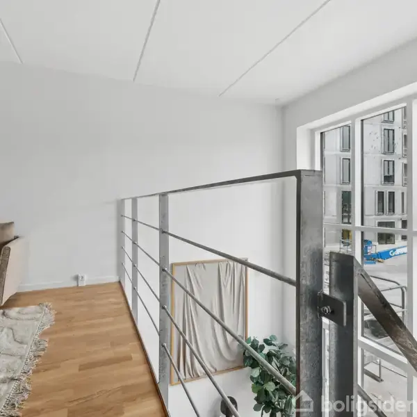Metalgelænder adskiller en hems med sofa og tæppe fra store vinduer, der vender ud mod en urban udsigt med moderne bygninger.