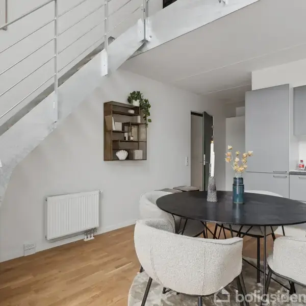 Et rundt spisebord med hvide stole står under en moderne trappe i en minimalistisk indrettet stue. Til venstre ses en radiator og en reol på væggen.