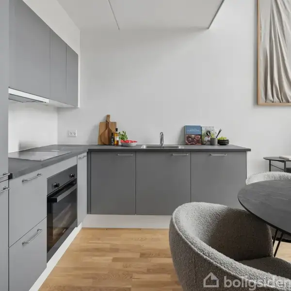 Gråt køkken med integrerede apparater, herunder ovn og kogeplade. Rundt spisebord med vase og stole. Lys træparketgulv og moderne kunst hængende på hvid væg.