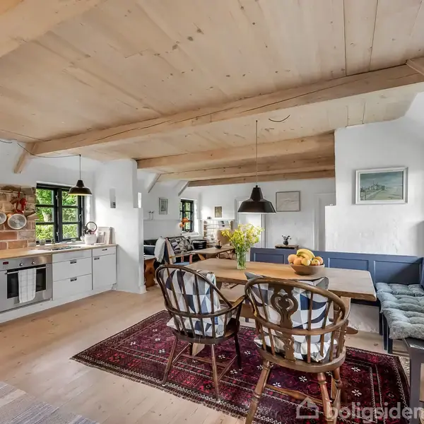 Køkkenalrum med træloft og lyse vægge. Rundbord med stole og bænk omkranset af vinduer. Køkken med komfur og krydderihylde. Blomster og frugt på bordet, tæppe på gulv.