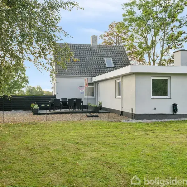 Hvidmalet hus med sort tag, omgivet af en grøn græsplæne. Terrasse med borde og stole ved siden af. Træer og hegn skaber en afgrænset have i et landligt område.