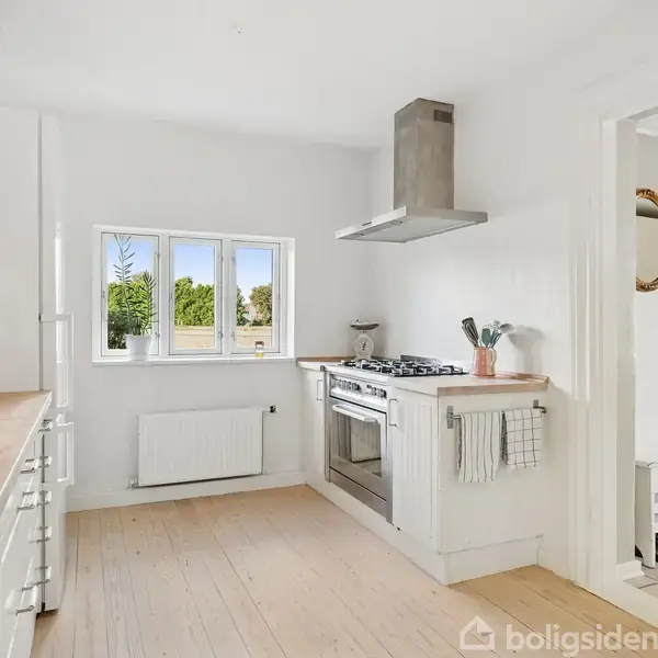 Køkken med hvidt interiør har en komfur med ovn og emhætte, ved siden af et vindue med udsigt til grønne træer. Bordpladerne er af lyst træ.