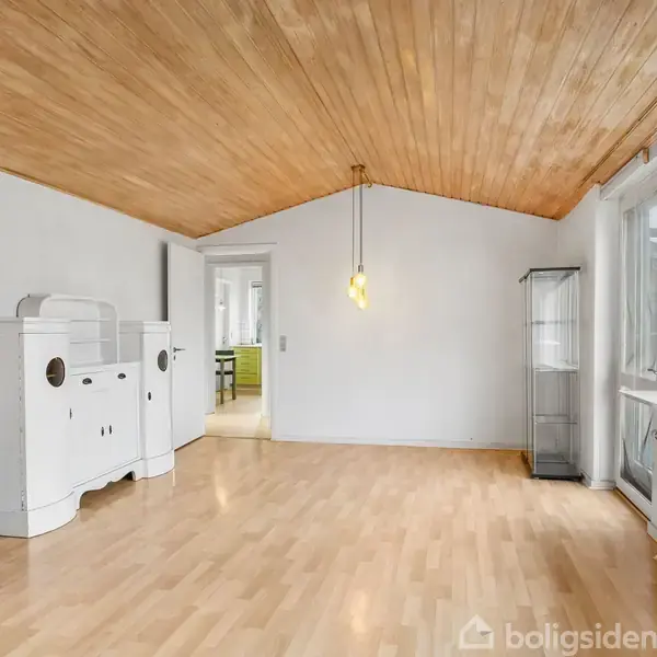 Et rum med lyst trægulv og loft, en hvid skænk og et glasvitrine i hjørnet. En gul lampe hænger fra loftet, og store vinduer til højre giver udsigt til haven.