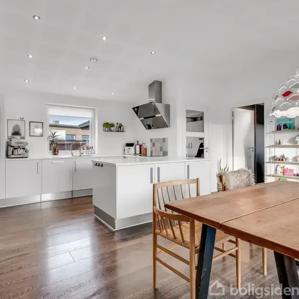 Køkken med hvidt interiør og åbne hylder, der står i forgrunden. Træbord og stole i spisestueområdet. Rummelig lys med lamper i loftet og planter ved vinduet.