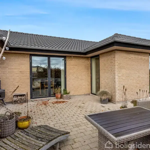 Terrasse med flisebelægning foran et hus med lyse mursten. Udendørs møbler og krukker med planter er placeret omkring området. Huset har store glasdøre og et mørkt tag.