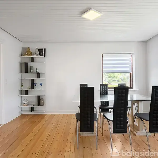 Spisebord med seks sorte stole står på lyst trægulv i et rent, lyst rum. Hylder til venstre bærer dekorative genstande. Rummet har to store vinduer med persienner.