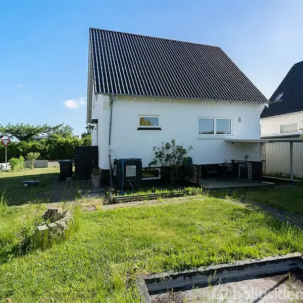 Hvidt hus med sort tag står på en græsplæne med bede. I baggrunden ses træer og buske under en blå himmel.