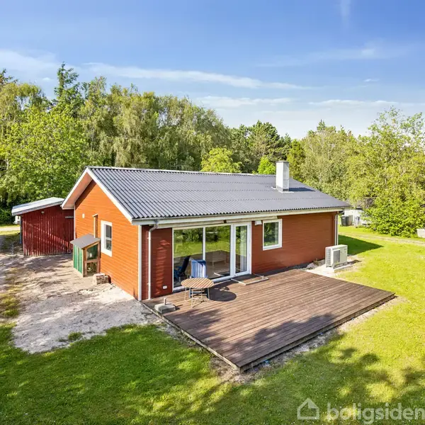 Rødt træhus med gråt tag står stille i en grøn have med frodige træer. En træterrasse med havemøbler ses foran huset. Grusvej løber langs siden.