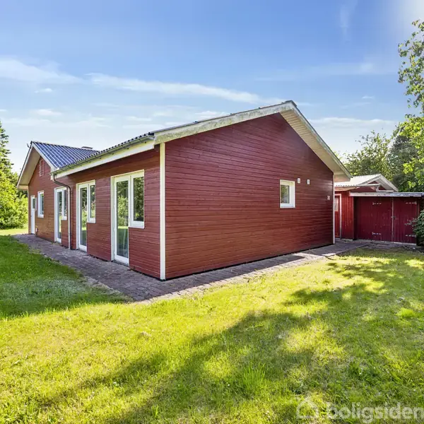 Rødt træhus står stille. Omgivet af frodige grønne områder og høje træer under en klar blå himmel.