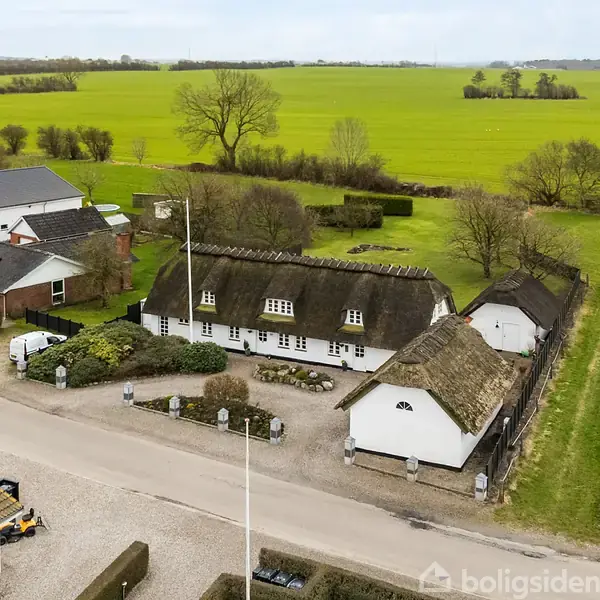 Bindingsværkshus med stråtag står stille langs en vej. Det er omgivet af flere huse og åbne marker i et landdistrikt. Strukturen står ud med sine traditionelle arkitektoniske træk.
