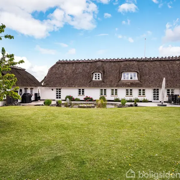 Stråtækt hus med hvid facade står i en frodig have omkranset af træer. Flere vinduer pryder facaden, og græsplænen strækker sig foran huset under en blå himmel.