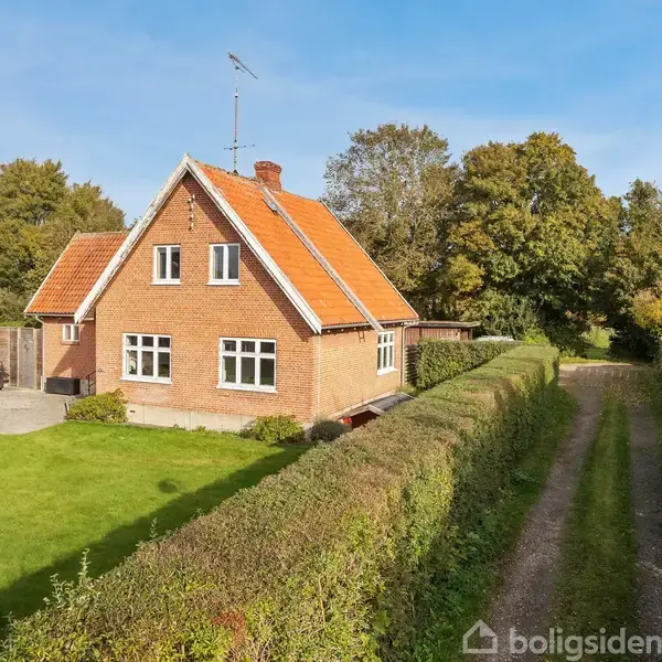 Et rødt murstenshus med rødt tag står ved siden af en grusvej. Huset er omgivet af en grøn have og hække, mens træer danner baggrund.