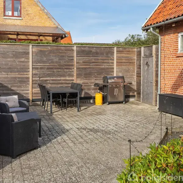 Terrassen har sorte lounge-møbler og grill, omgivet af høje træhegn. Til højre ses en murstensvæg med husnummeret 22. I baggrunden er et andet hus med gult murværk.