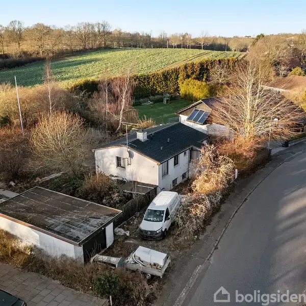 Et hvidt hus med en varevogn parkeret foran ses fra luften. Det er omgivet af buske og træer, med en landevej til højre og marker i baggrunden.