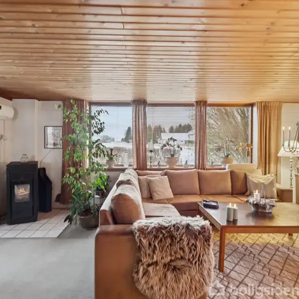 Stue med hjørnesofa dækket med puder, står ved store vinduer. I hjørnet er en opvarmningselement ved siden af en plante. Et stort træbord med levende lys i midten skaber hygge.