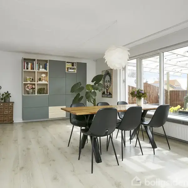 En spisestue med et træbord omgivet af seks sorte stole i midten. Store vinduer slipper lys ind og viser udsigt til haven; en bogreol og planter pynter rummet.