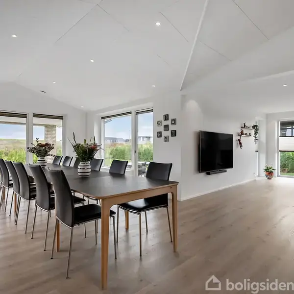 Et moderne spisebord med otte stole omgivet af et lyst, rummeligt opholdsrum. Store vinduer giver udsigt til en grøn have. En vægmonteret fladskærm tilføjer en moderne atmosfære.