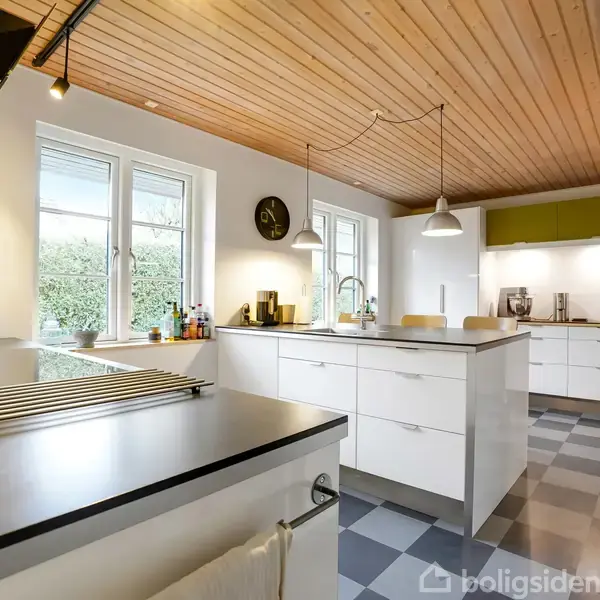 Køkken med hvide skabe og en central ø, oplyst af hængende lamper, i en rummelig og lys atmosfære. Træloft og skakternet gulv tilføjer varme til det moderne design. Krydderiglas ved vinduet.