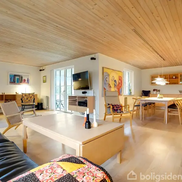 Stue med trægulv og træloft. En sort lædersofa og et lyst sofabord står i forgrunden, mens et spisebord og stole ses i baggrunden. Vægmalerier og en TV-skærm på væggen.