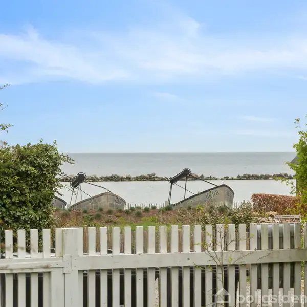 Hvid træport står åben mod en kystlinje med fiskerbåde i nærheden. Grønt buskads omgiver området, og havet strækker sig under en blå og skyet himmel.